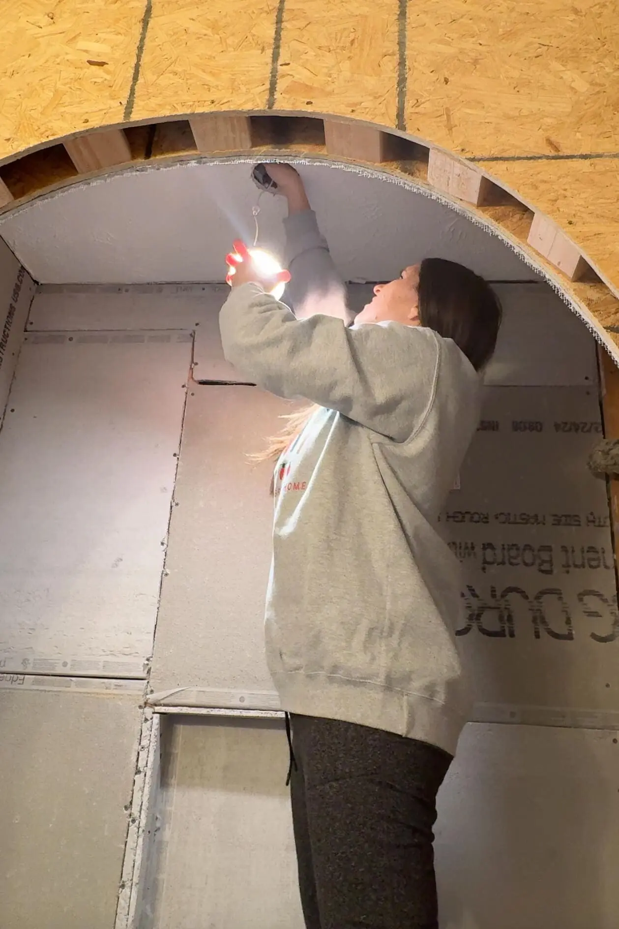 Woman in a gray sweatshirt installing a light fixture inside a shower during a DIY renovation project, with exposed wood framing above.